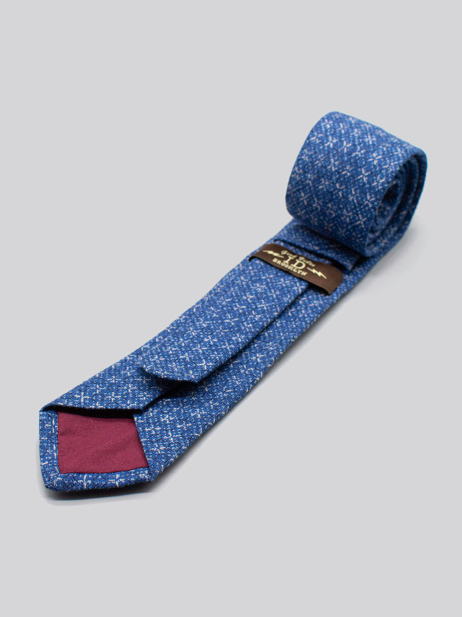The Japanese Printed Blue Kross Tie by ID Menswear, handmade in Brooklyn from 100% cotton, features a maroon tip and visible label. Rolled and textured, it’s displayed on a light gray background.