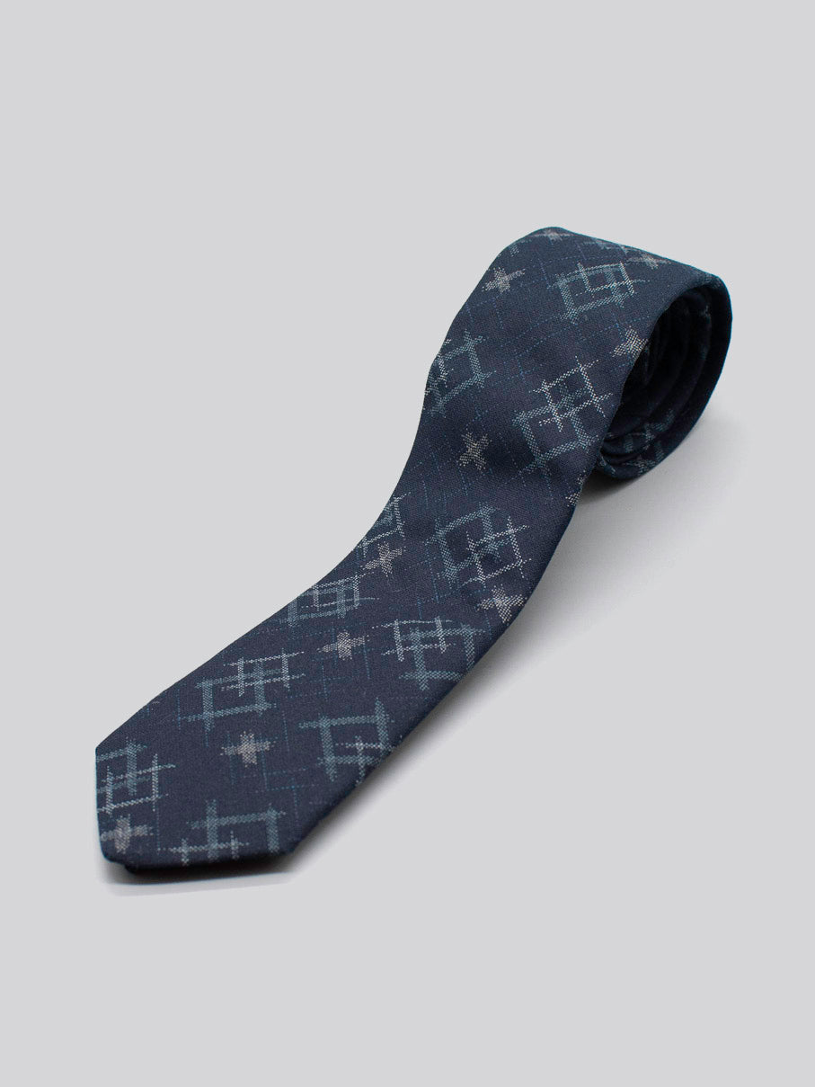 The Japanese Printed Ikat Tie by ID Menswear features a navy blue geometric print in lighter blue and gray, handmade in Brooklyn from 100% cotton, and is shown rolled up on a light gray background.