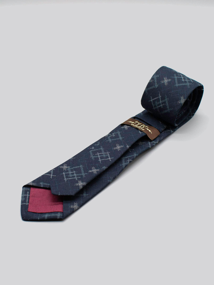 The Japanese Printed Ikat Tie by ID Menswear is a handmade navy tie with a subtle geometric pattern, crafted in Brooklyn from 100% cotton. It has a maroon lining, brown tag, and is shown on a plain light gray background.