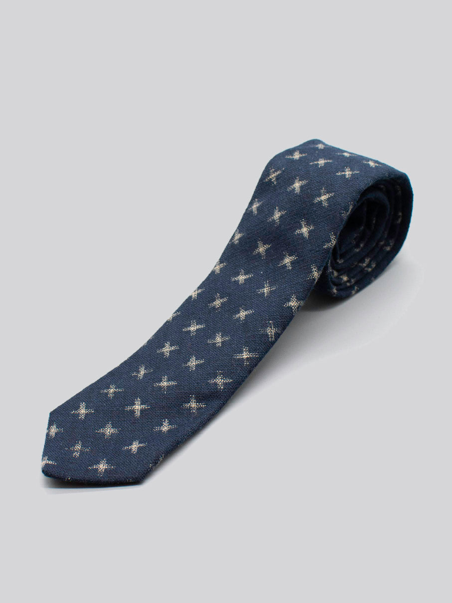 The Japanese Printed Kross Tie by ID Menswear is a navy blue tie handmade from 100% cotton, featuring a subtle pattern of small light crosses, and is shown neatly rolled on a plain light gray background.