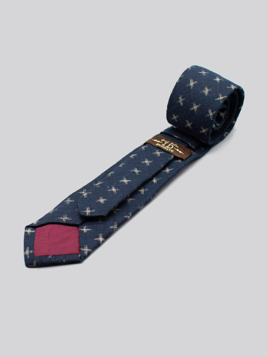 The Japanese Printed Kross Tie by ID Menswear is a rolled navy blue tie with white star prints, handmade from 100% cotton, and features a maroon accent on its inner tip, shown against a light gray background.