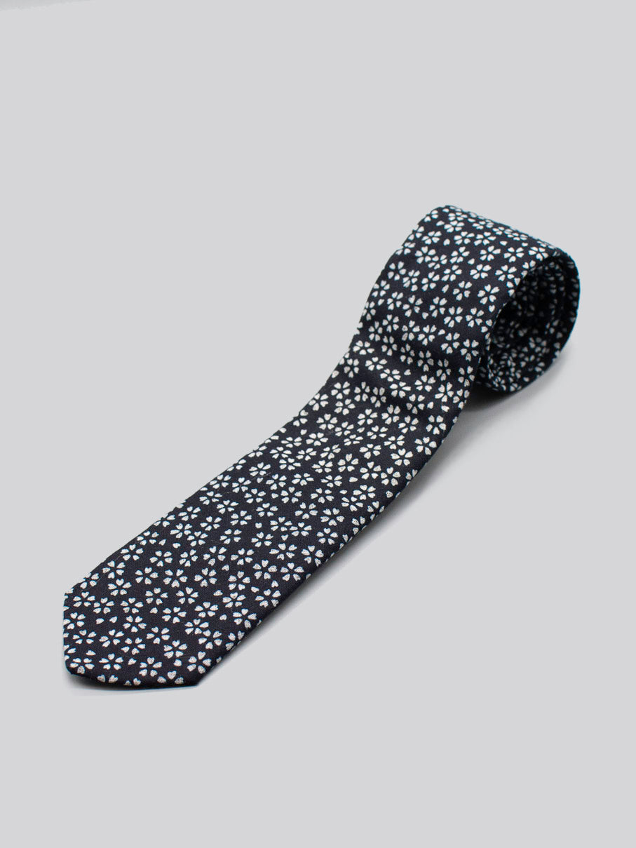 The Japanese Printed Mini Bloom Tie by ID Menswear is a black 100% cotton tie with a white floral pattern, handmade in Brooklyn from Japanese print fabric, shown rolled up on a plain light gray background.