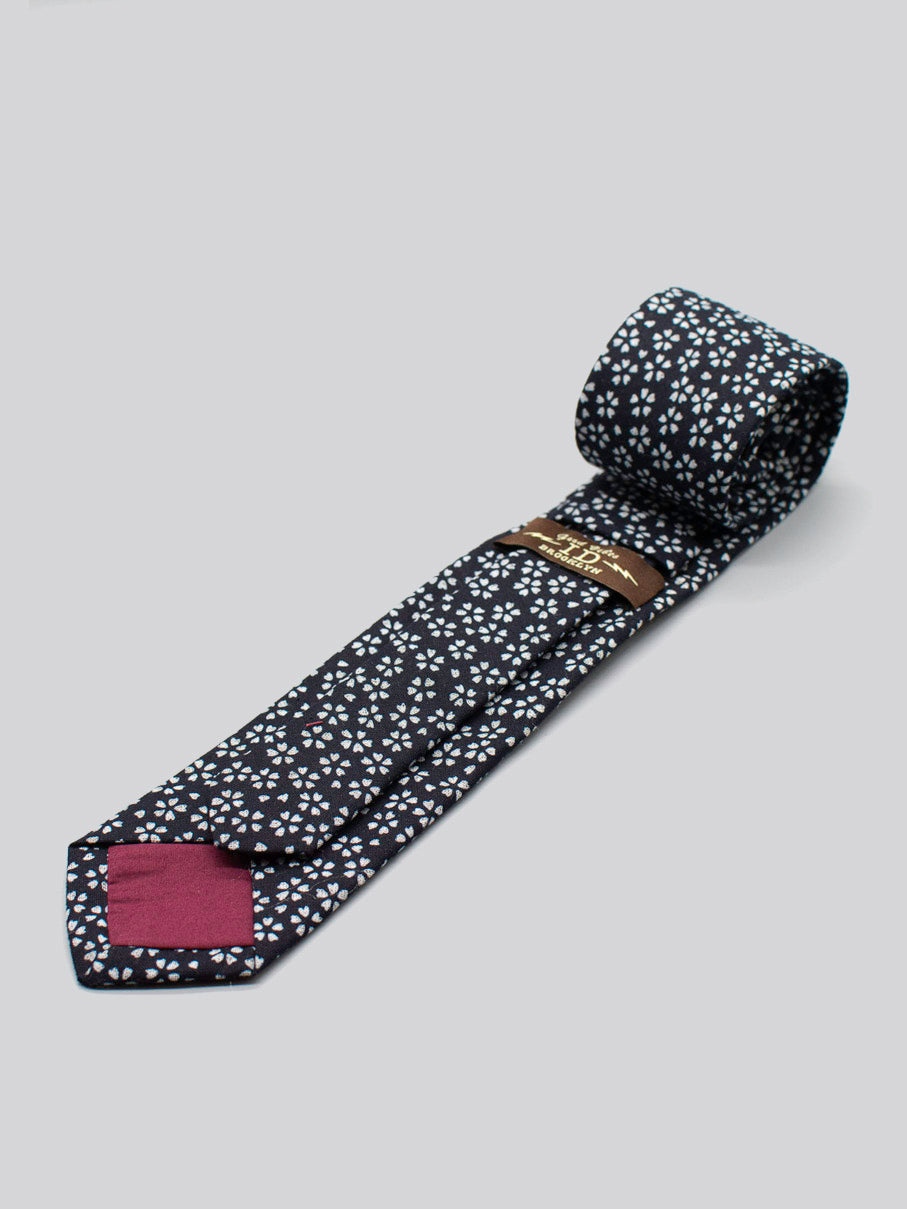 The Japanese Printed Mini Bloom Tie by ID Menswear is handmade in Brooklyn from 100% cotton, featuring a black base with white floral print, maroon accent, and brown label—all displayed on a light gray background.