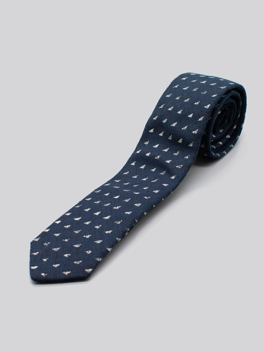The Japanese Printed Nara Tie by ID Menswear is a navy blue cotton tie handmade in Brooklyn, featuring a subtle white sailboat pattern. One end is rolled and displayed on a light gray background.