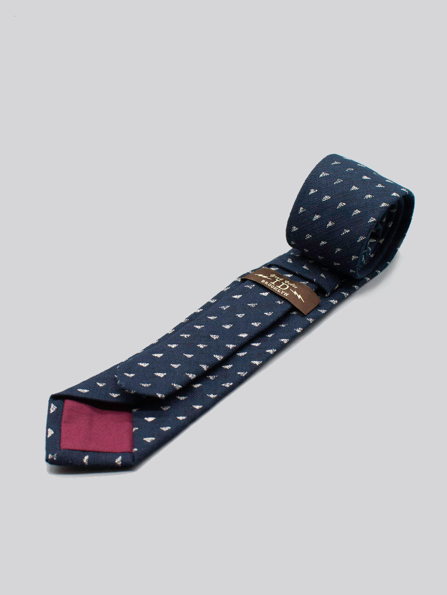 The Japanese Printed Nara Tie by ID Menswear is a navy tie with a small white pattern, handmade in Brooklyn from soft cotton. It has a maroon tip detail and brown brand label underneath, shown on a light gray background.