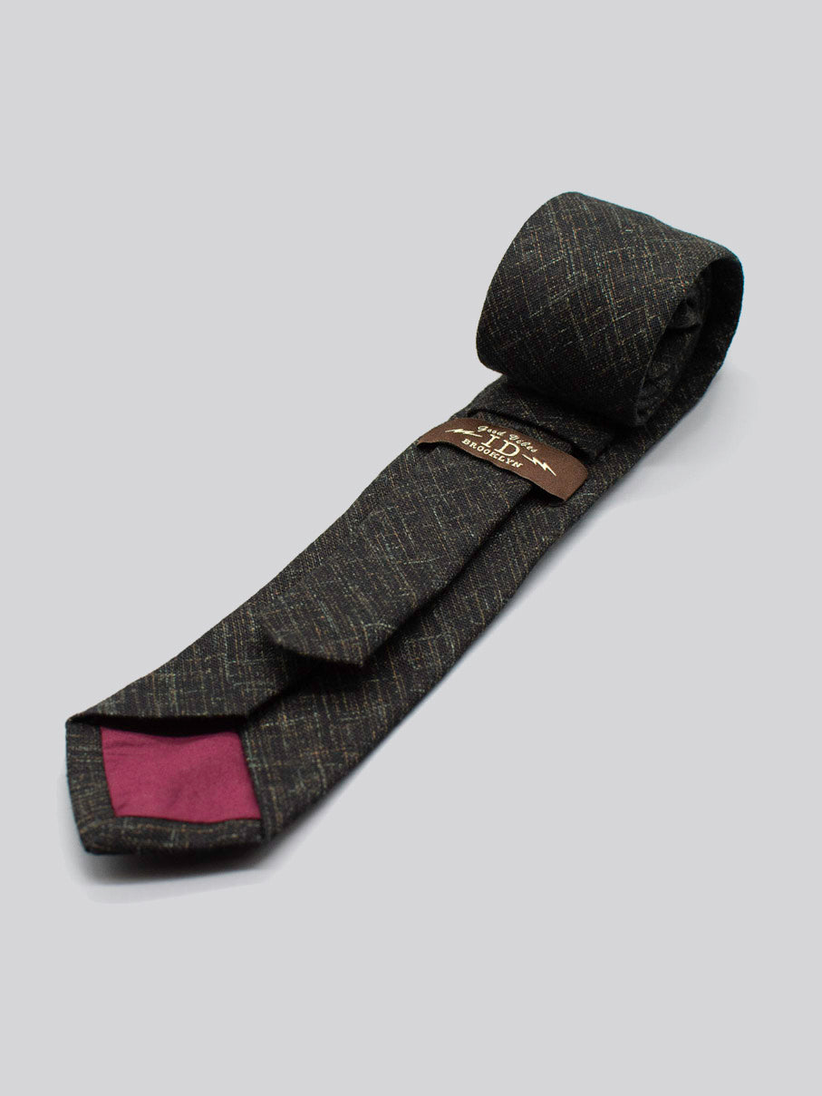 The ID Menswear Japanese Printed Ninja Tie is handmade in Brooklyn from 100% cotton, with a dark textured crisscross pattern, maroon inner lining, and brown label with gold text—shown rolled on a light gray background.
