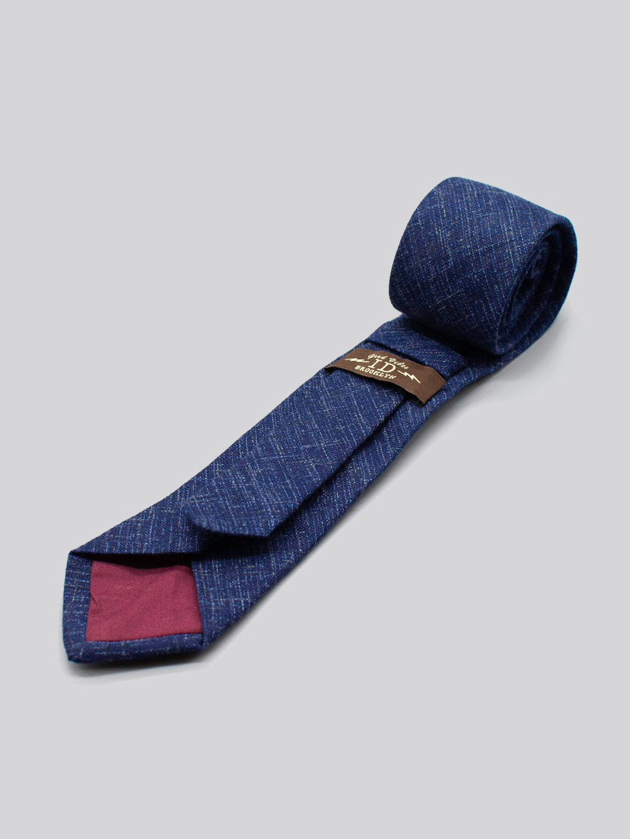 The Japanese Printed Samurai Tie by ID Menswear is a blue textured tie with a subtle pattern, handmade from 100% cotton. It features a maroon-lined inner tip and brown label, shown rolled on a light gray background.