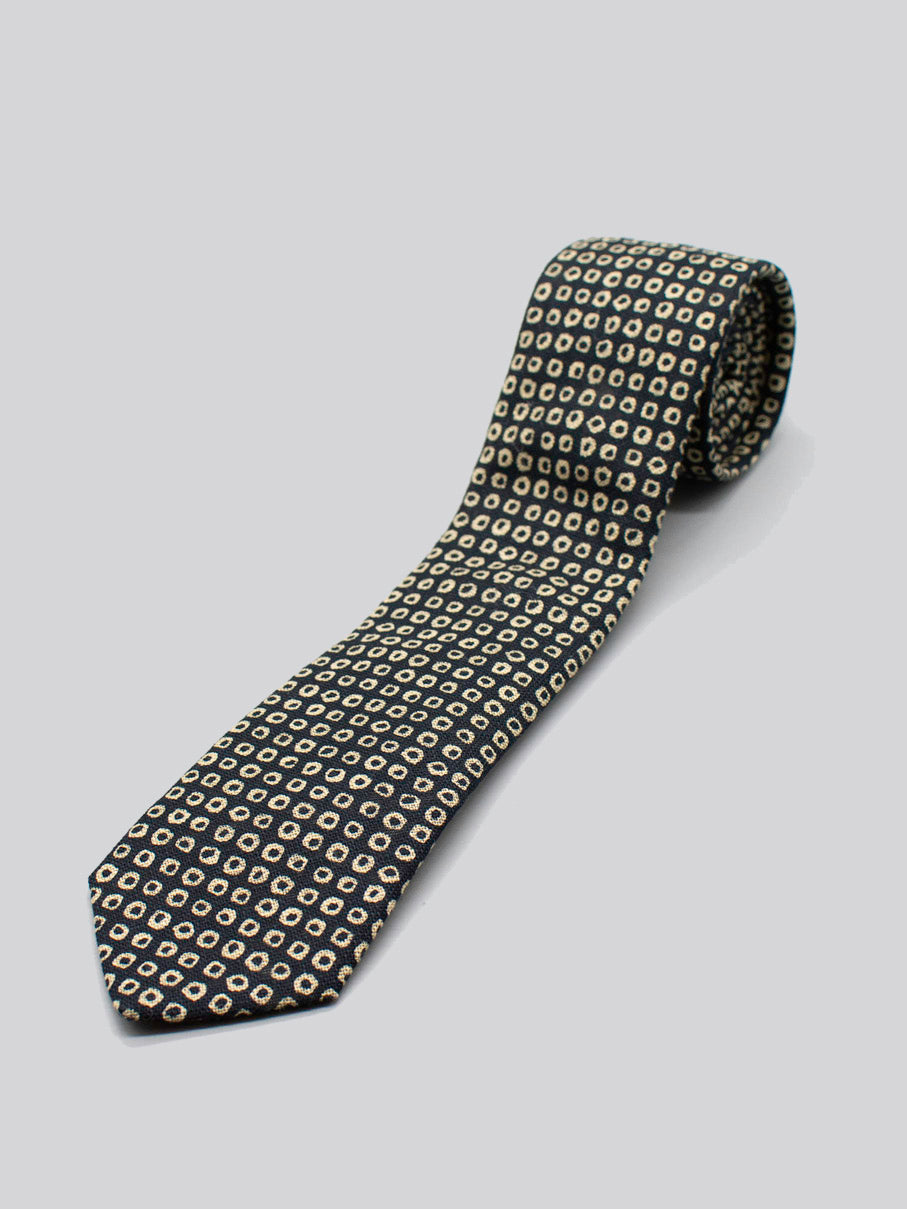 The Japanese Printed Shibori Tie by ID Menswear, handmade in Brooklyn from Japanese-printed cotton, showcases a black background with small yellow and white squares, and is displayed rolled up on a light gray surface.