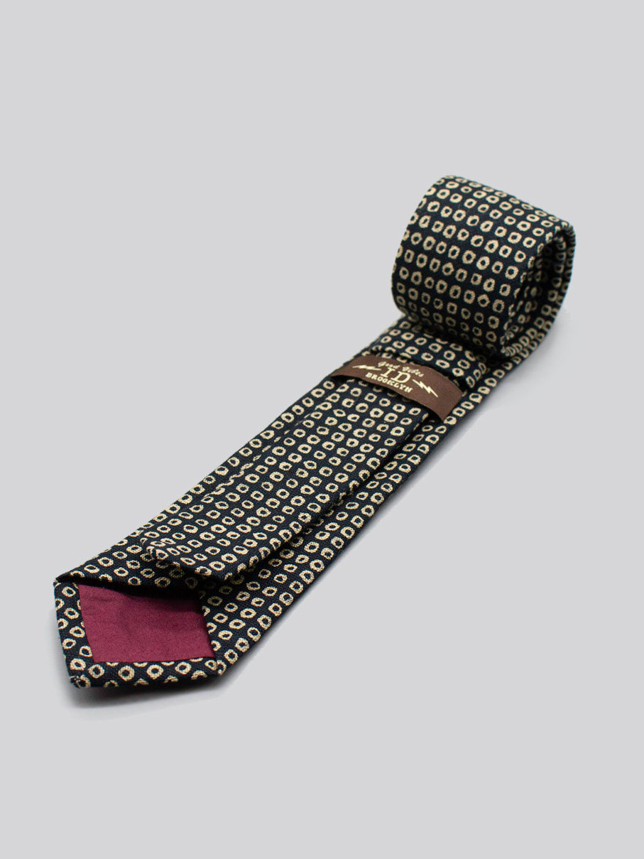 The Japanese Printed Shibori Tie by ID Menswear features a dark cotton base with small light geometric patterns, a maroon lining, and label. Handmade in Brooklyn from Japanese-printed cotton, its shown rolled and flat on a gray background.