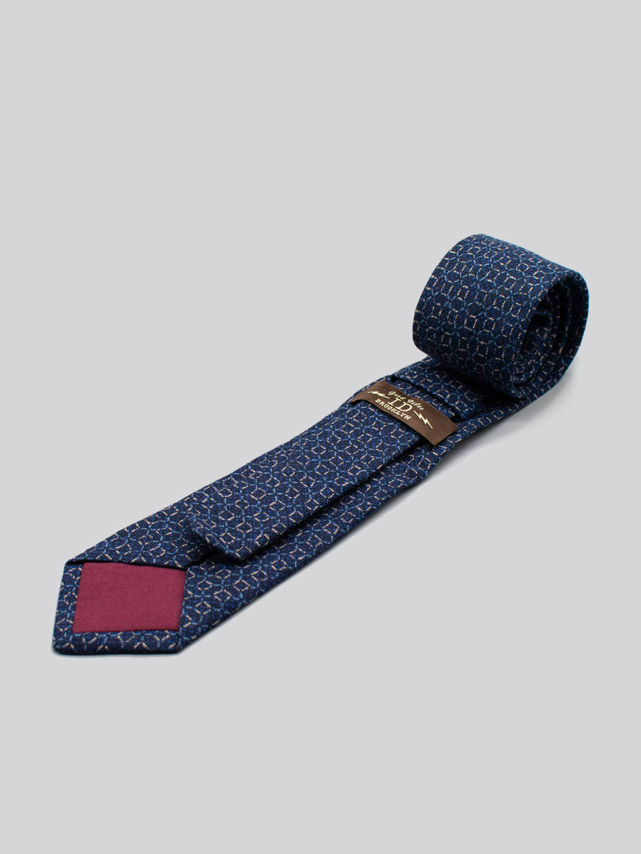 The Japanese Printed Web Tie by ID Menswear features a subtle blue geometric pattern, handmade from 100% cotton. It has a burgundy tip inset, brown back label, and is shown rolled on a light gray background.