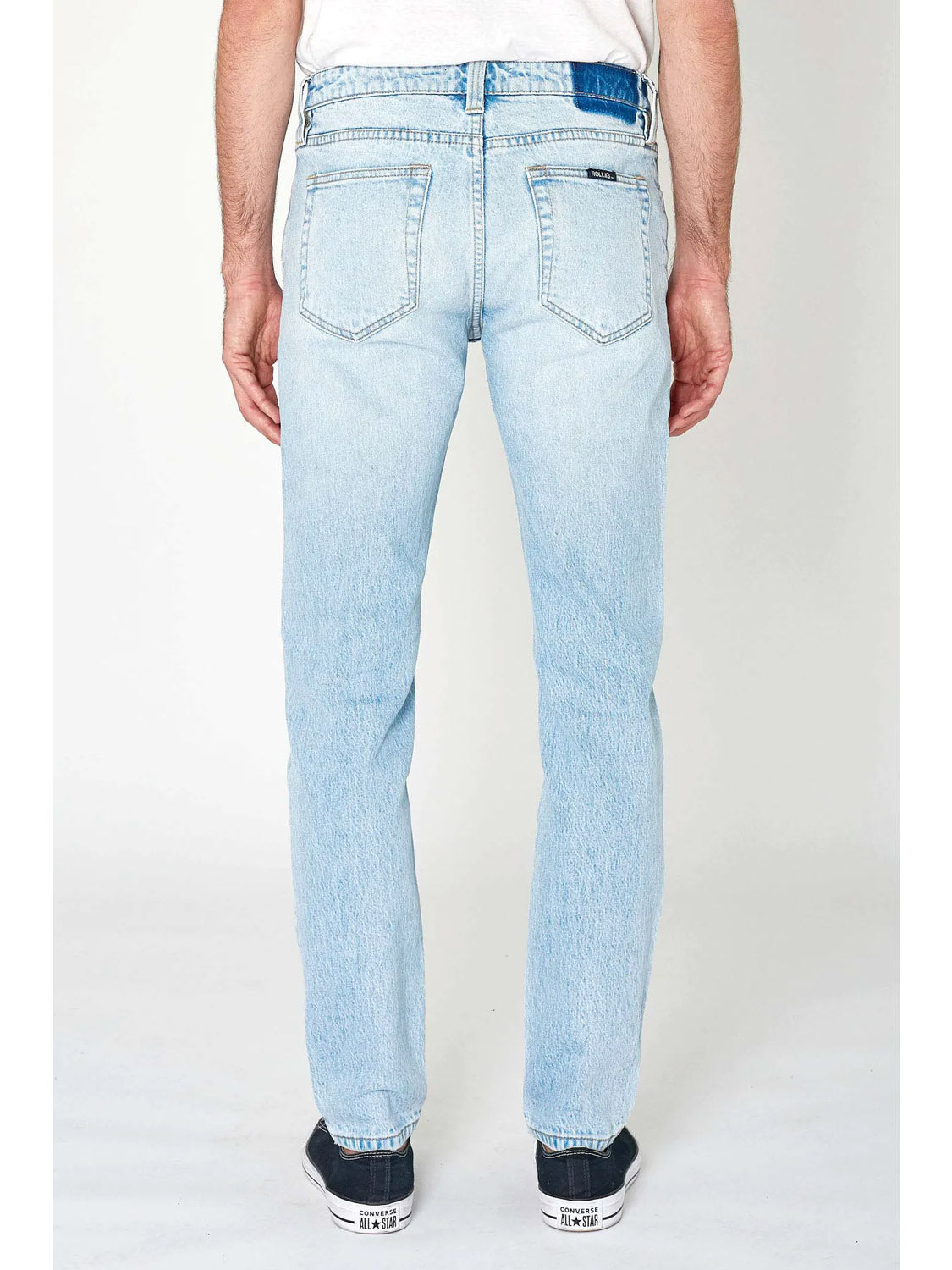 A person is wearing Rollas Tim Slims Bonzer Blue jeans, a white shirt, and black sneakers, standing with their back to the camera against a plain white background.