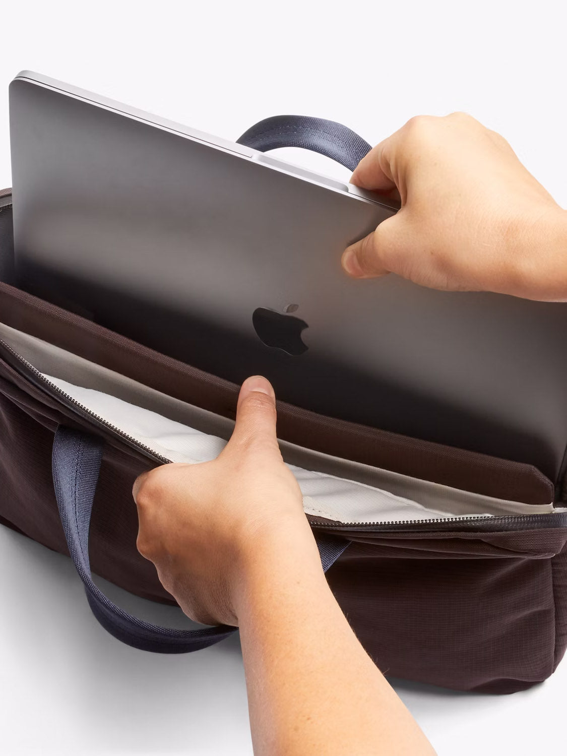 A person puts a silver laptop into the padded 16” laptop sleeve of the brown Bellroy Tokyo Work Bag - 20L, which features dark handles.