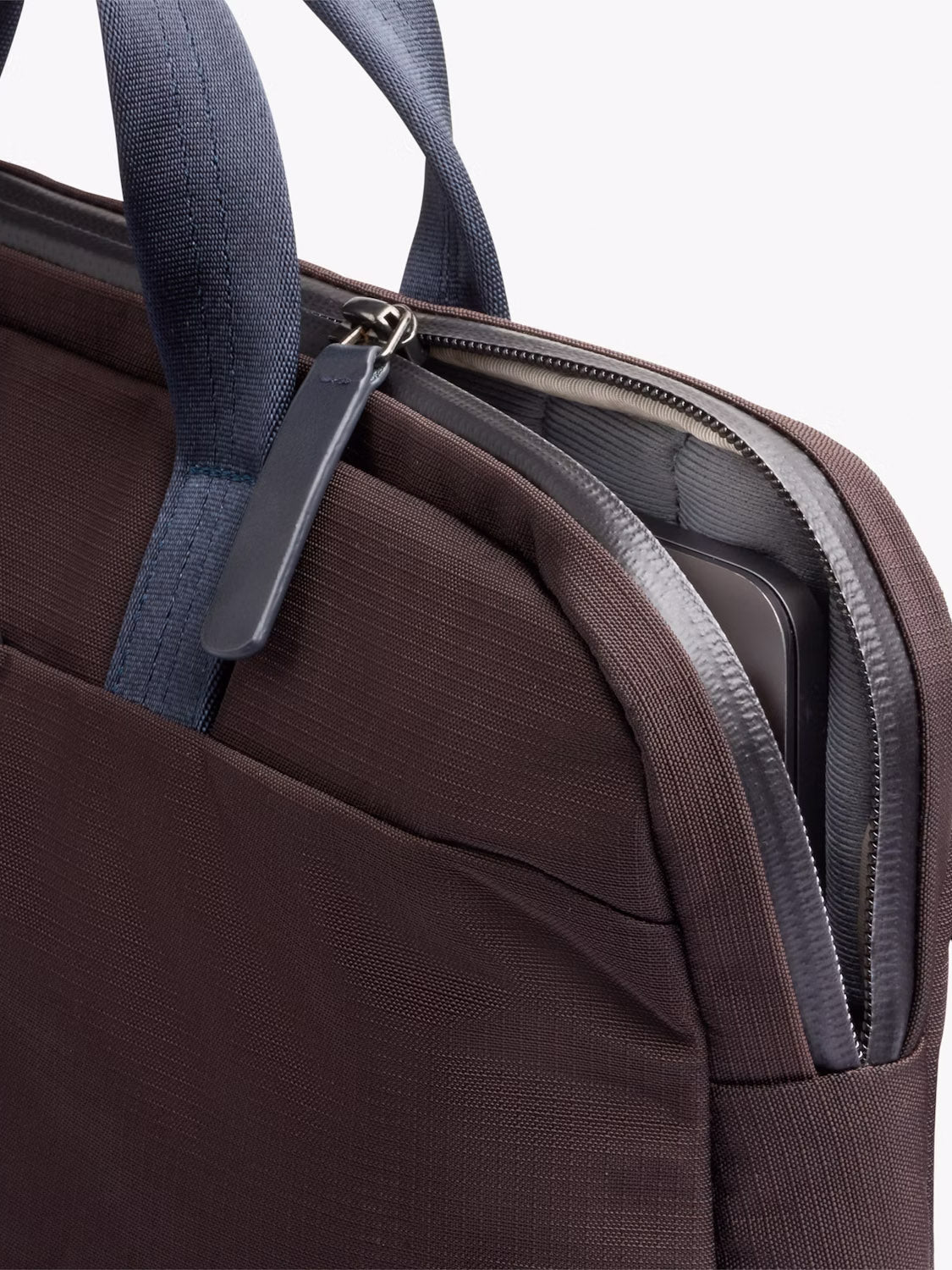 A close-up of the Bellroy Tokyo Work Bag - 20L in brown, featuring dark blue handles and a visible 16” laptop sleeve. The textured fabric and zipper stand out, with the bag partially unzipped against a light background.