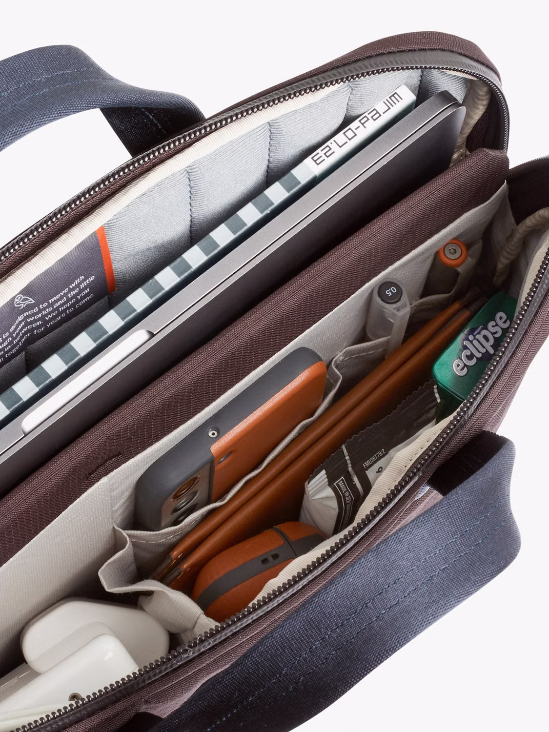 The Bellroy Tokyo Work Bag - 20L, in brown and gray, features organized compartments to hold a laptop, notebook, phone, orange wallet, pack of Eclipse gum, AirPods case, pens, and a 16” sleeve for easy on-the-go organization.
