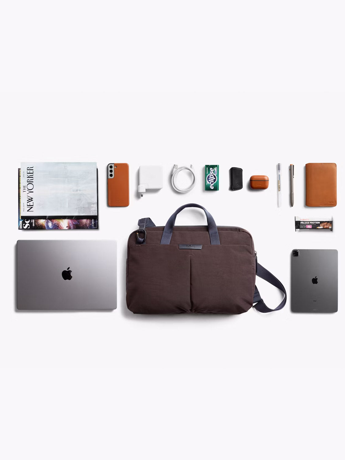 A neatly organized flat lay displays the Bellroy Tokyo Work Bag - 20L in brown, alongside a closed MacBook, iPad, magazines, phone, wallet, charger, cables, AirPods, gum, keys, and a chocolate bar on a white background.