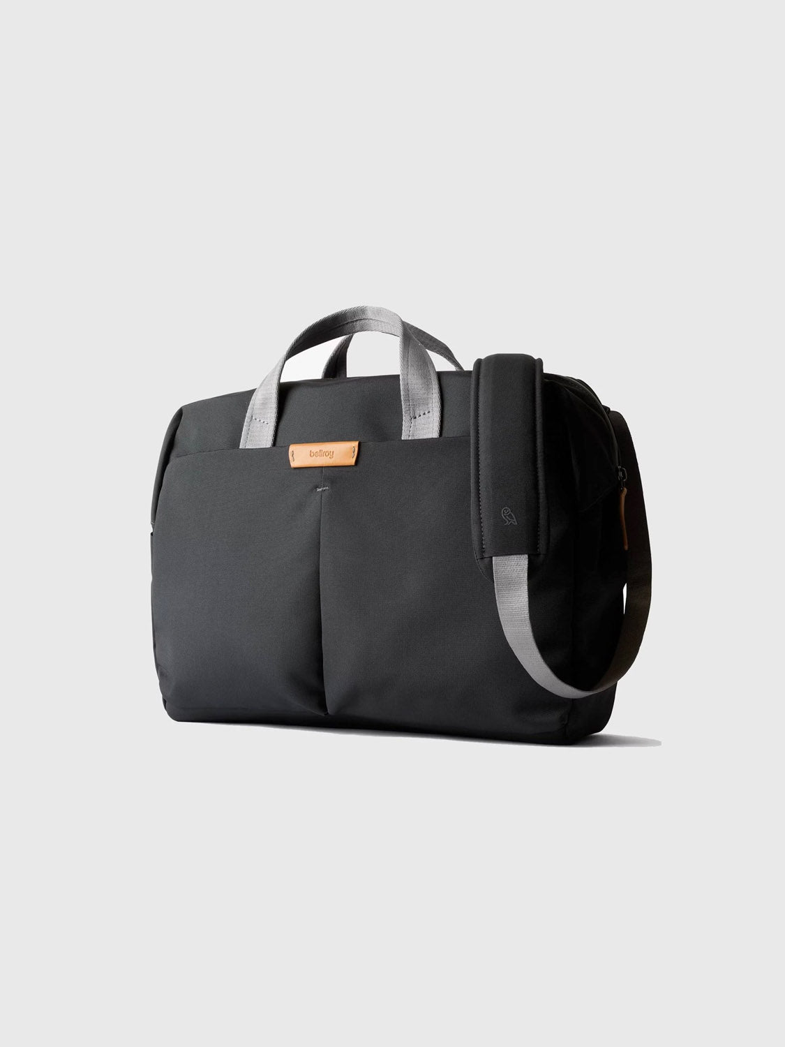 The Bellroy Tokyo Work Bag - 20L is a black fabric duffel with gray handles, tan leather accent, and gray shoulder strap, ideal for commuting with a 16" laptop sleeve—shown against a plain light gray background.