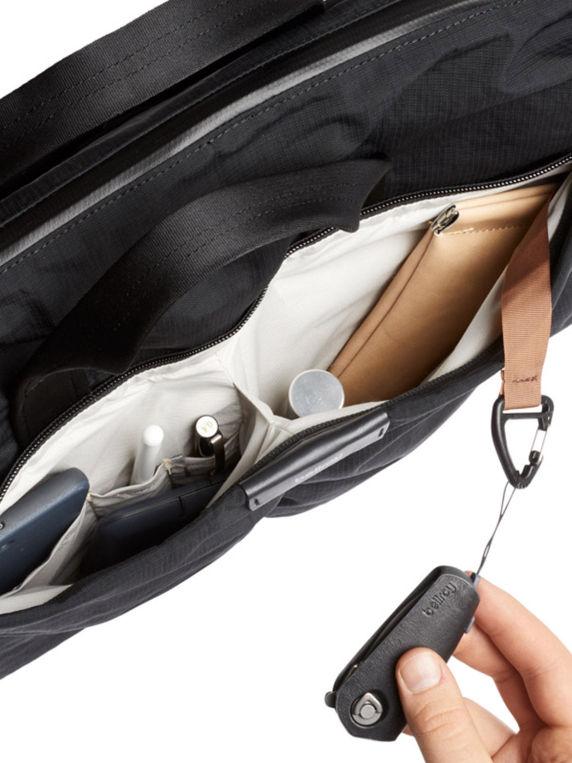 A hand pulls a retractable key holder inside the Bellroy Tokyo Work Bag - 20L, a black commuter bag with a 16” laptop sleeve and organized compartments for pens, notebook, phone, and a brown case.