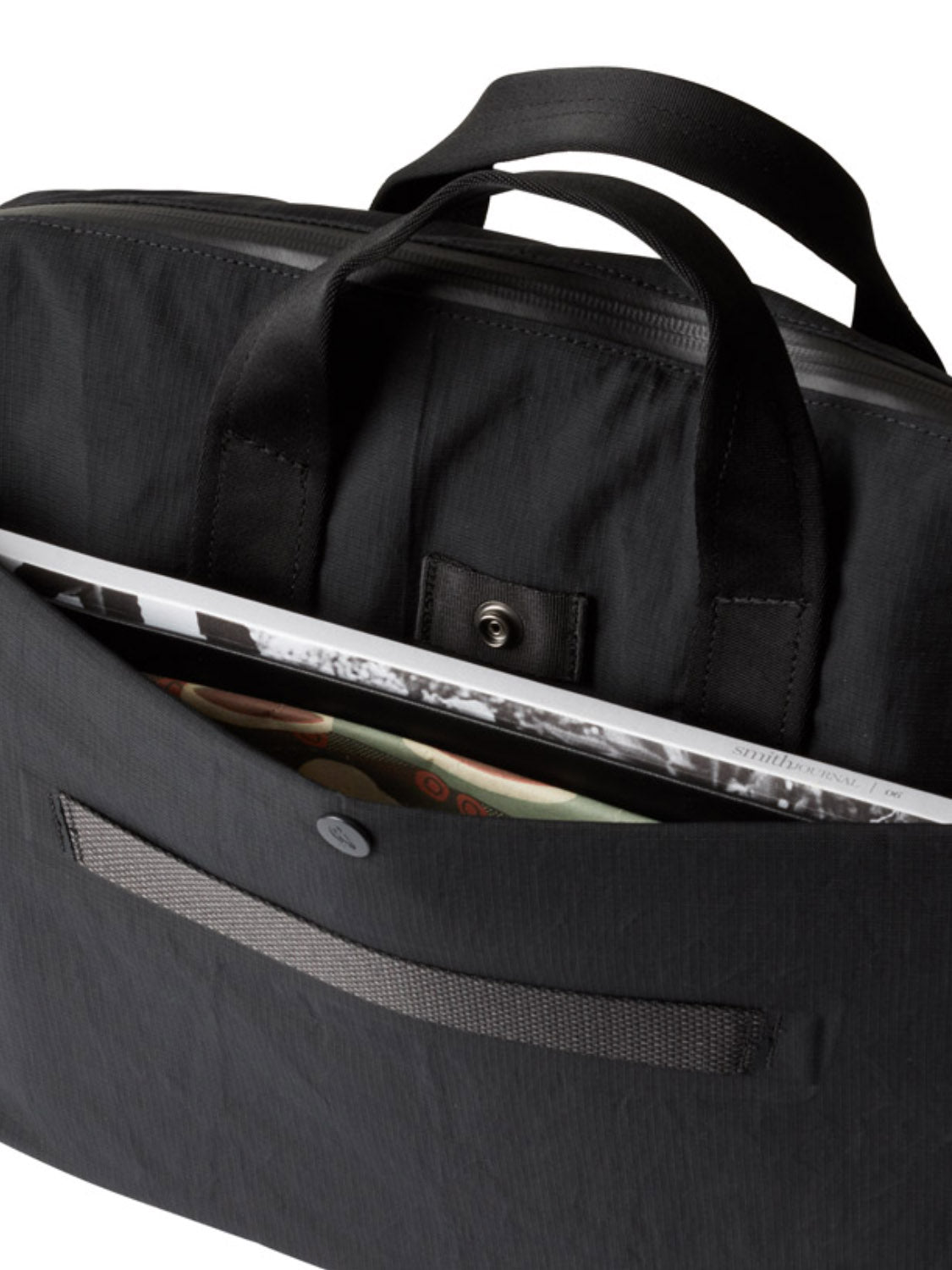 A close-up of the Bellroy Tokyo Work Bag - 20L, a black commuter bag with short handles, shows a magazine in the front pocket and buttoned flap. Inside, it features a 16" laptop sleeve for convenience.