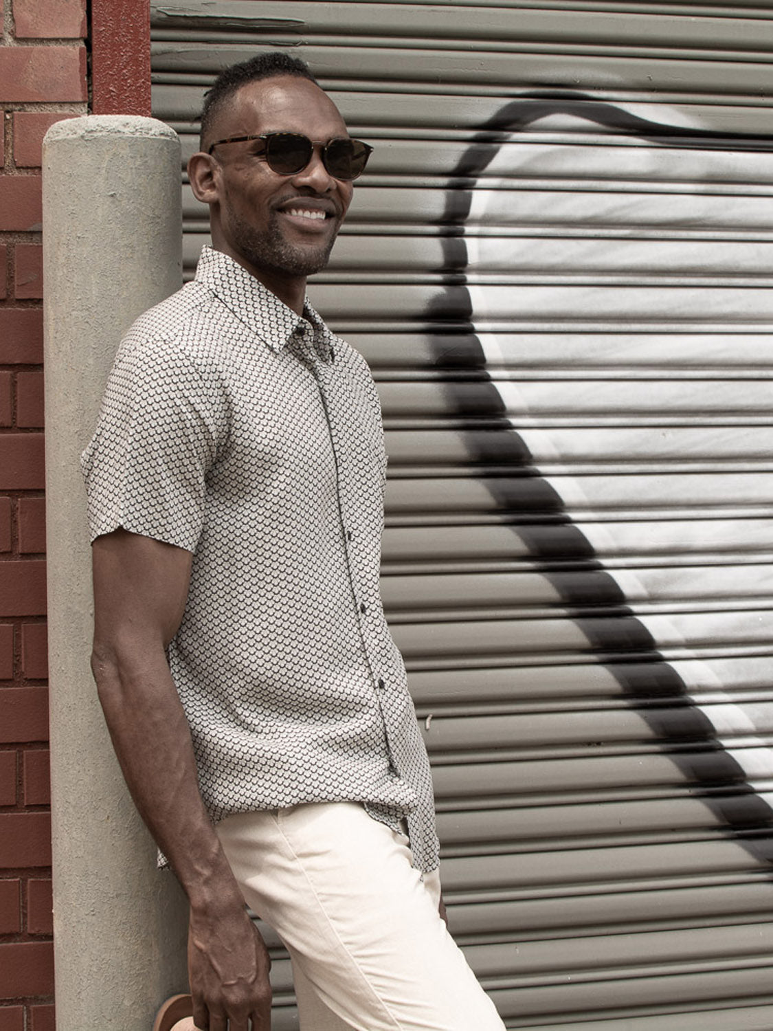A man in the Toyo Japanese Print Shirt by ID Menswear and sunglasses leans against a pole, smiling, with a large heart painted on a metal shutter behind him—perfect for warm weather style.