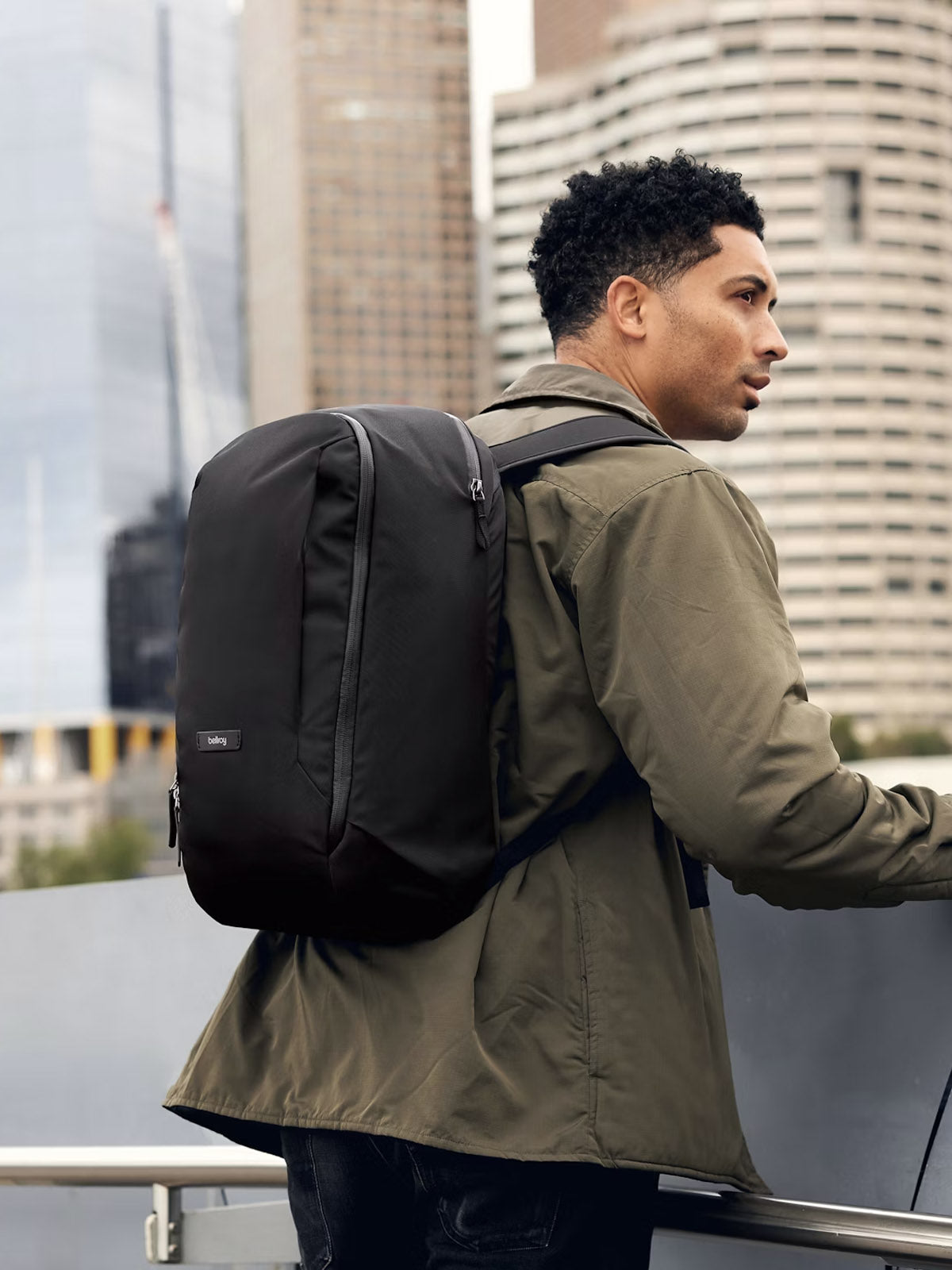 A man stands outdoors with the Bellroy Transit Workpack - 20L, a minimalist green jacket, and a spacious black backpack featuring a laptop sleeve. Modern city skyscrapers rise in the background.