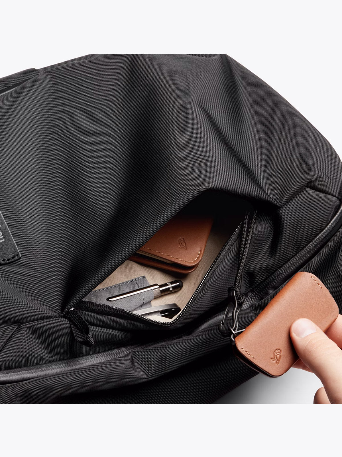 A hand opens the front pocket of the Bellroy Transit Workpack - 20L, revealing neatly organized items inside, such as a small brown leather wallet, a pen, pencil, and notepad.