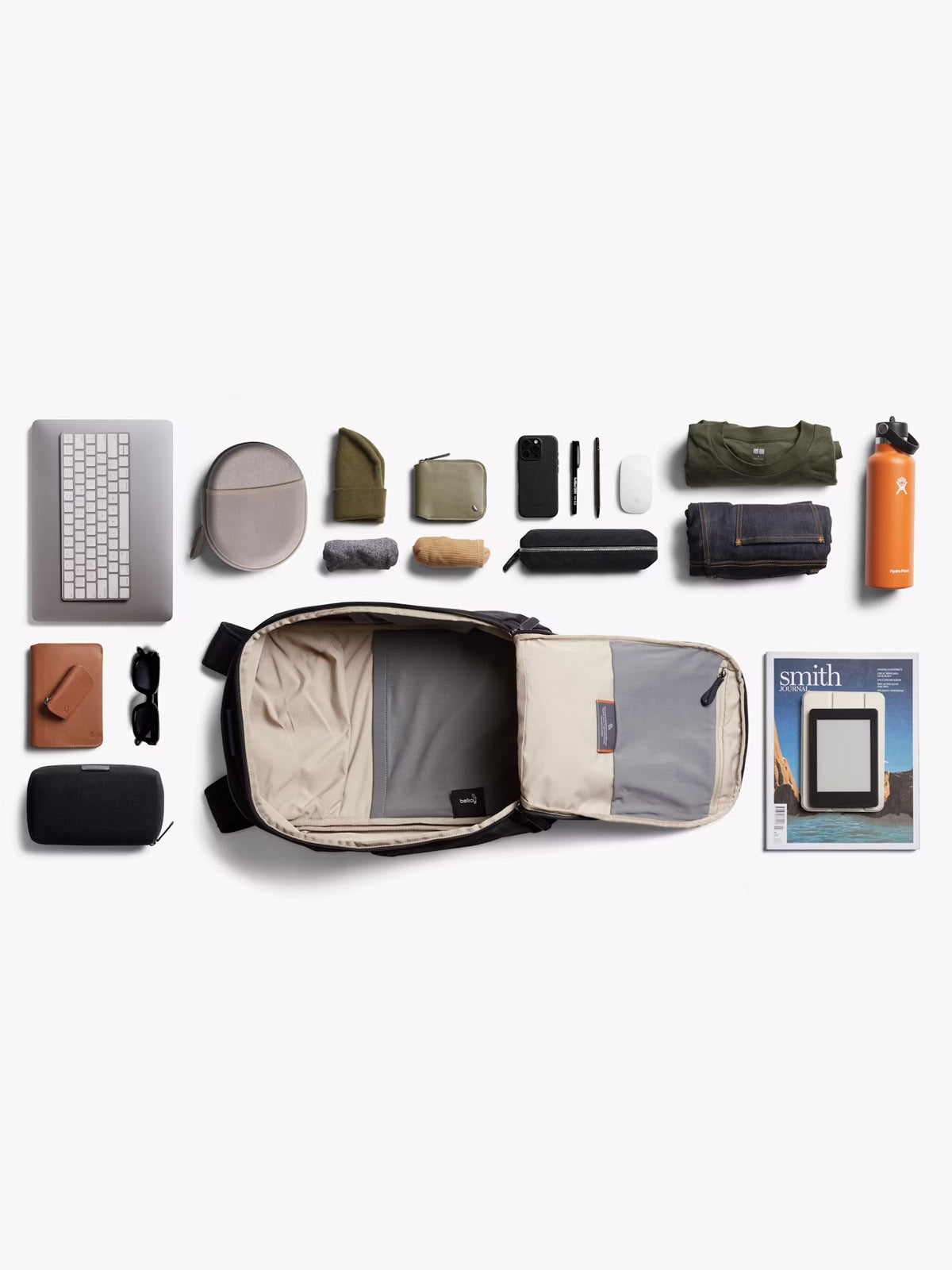 A neatly arranged flat lay of travel essentials, featuring the Bellroy Transit Workpack - 20L, laptop with a sleeve, keyboard, sunglasses, water bottle, minimalist clothing, pouches, notebook, pens, magazine and small tech accessories on a white background.