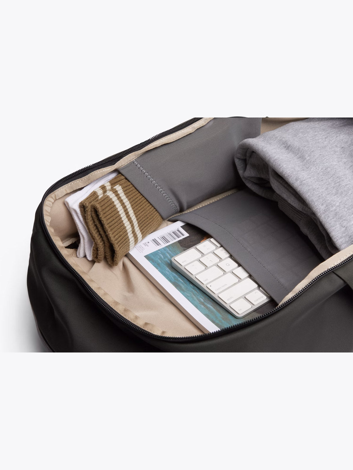 The Bellroy Transit Workpack - 20L is open, revealing neatly packed clothes, brown and white striped socks, a white keyboard, an ocean-themed magazine, and a minimalist laptop sleeve in the interior pocket.