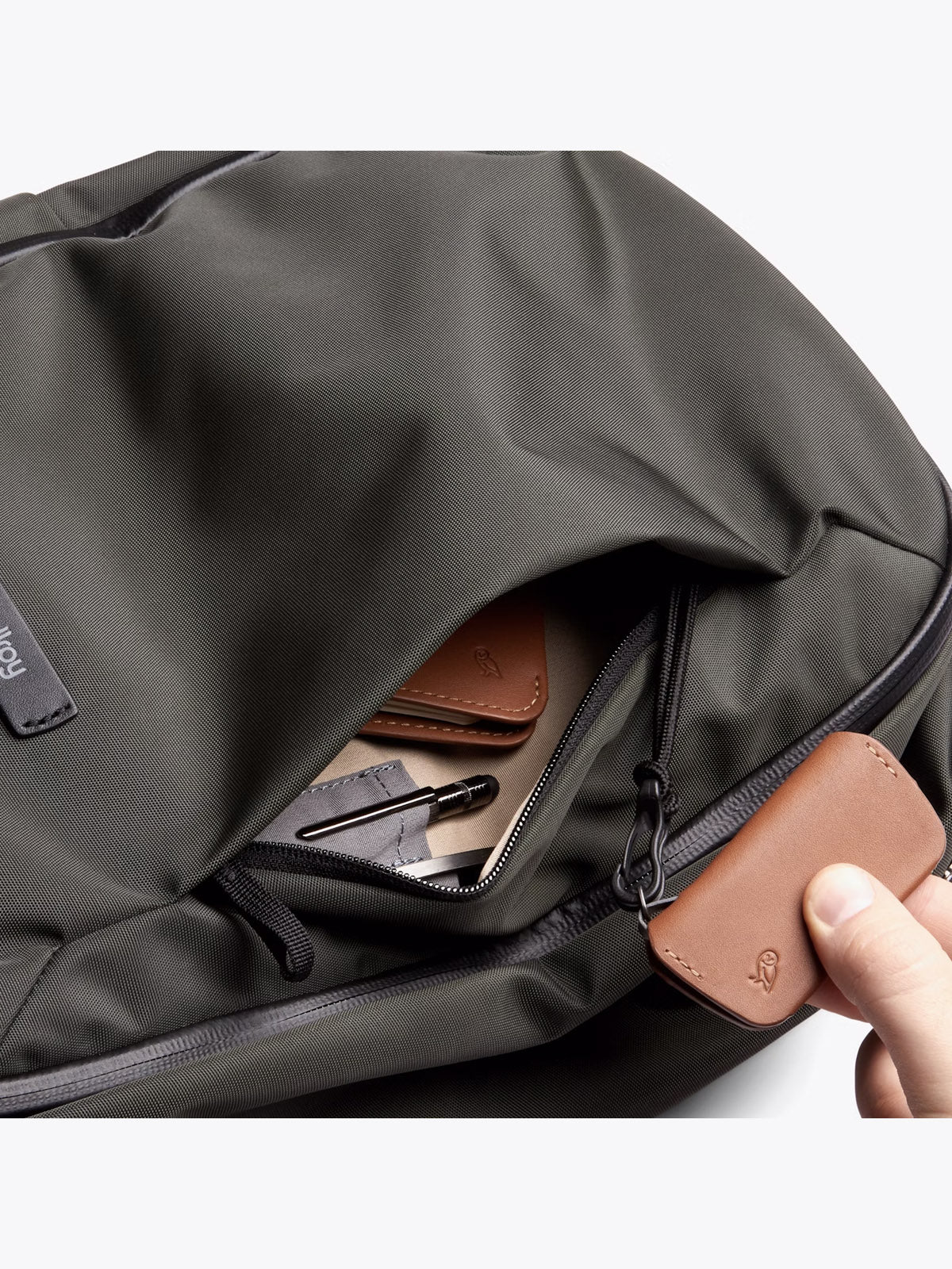 A hand opens the front pocket of the Bellroy Transit Workpack - 20L in dark gray, revealing neatly organized tan leather accessories, a laptop sleeve, and a black pen inside.