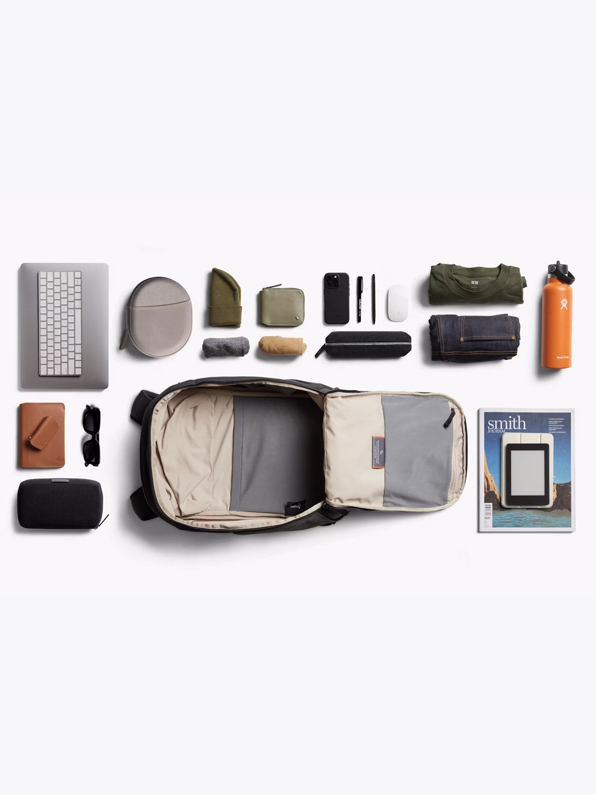 A minimalist flat lay features the Bellroy Transit Workpack - 20L, a laptop sleeve, keyboard, water bottle, sunglasses, clothes, pouches, pens, AirPods, wallet, and a magazine arranged on a white background.