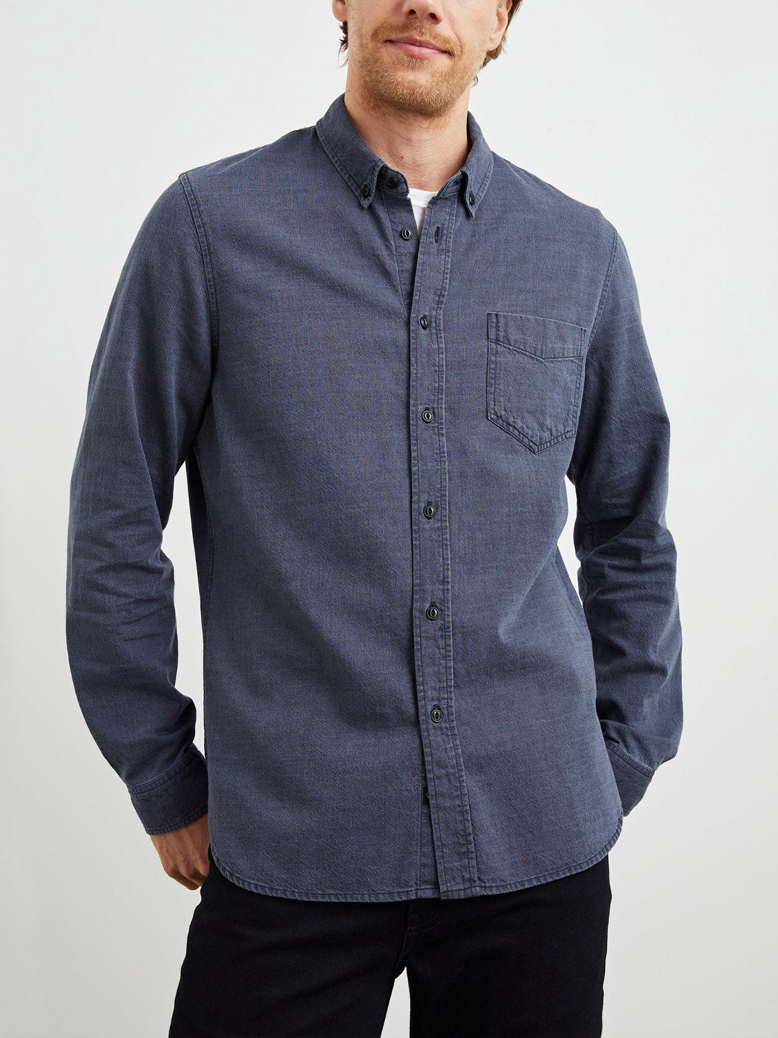 Wearing the Rails Tristan Shirt, a blue denim button-up made of 100% cotton with a front pocket, an individual with light hair stands against a plain white background. The shirt is untucked for a relaxed style, paired with black pants for effortless simplicity.