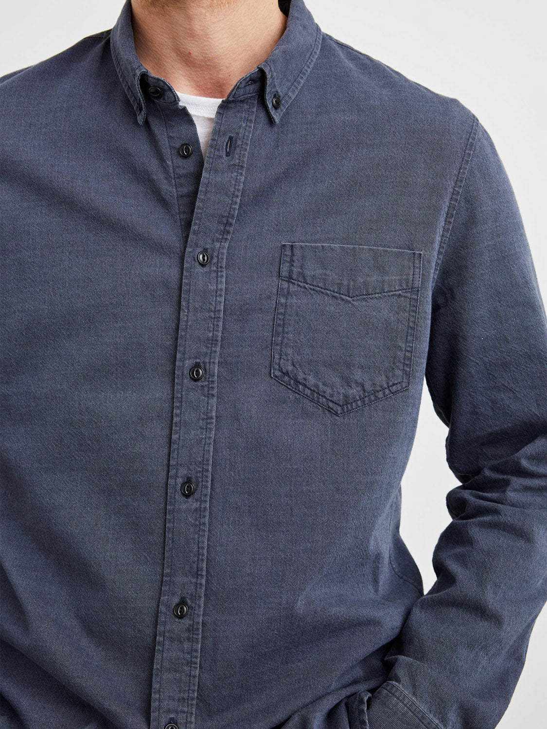 A person is wearing the Rails Tristan Shirt, a charcoal gray slub fabric button-up made of 100% cotton with black buttons and a chest pocket. They pair it over a white top, casually resting their left hand in their pant pocket against a plain white background.