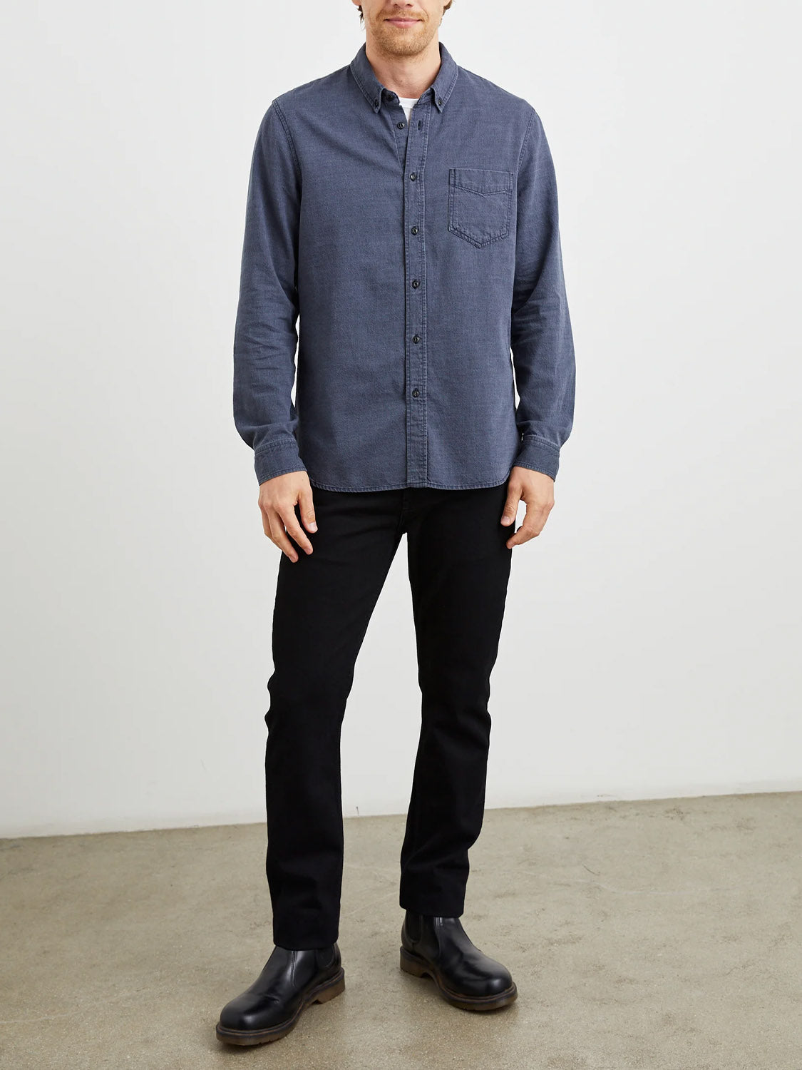 Tristan wears a Rails Tristan Shirt in blue, made from 100% cotton slub fabric, paired with black pants and boots. He stands against a plain white background, his hands resting relaxed by his sides on the simple concrete floor.