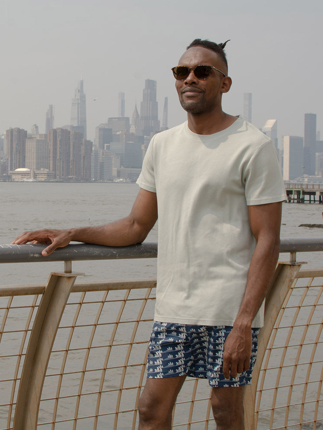 A man stands by a railing with a city skyline and water behind him, wearing sunglasses, a light T-shirt, and ID Menswears Tsunami Fast Dry 4-Way Stretch Swim Trunk made from recycled plastic bottles.
