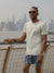 A man stands by a railing with a city skyline and water behind him, wearing sunglasses, a light T-shirt, and ID Menswears Tsunami Fast Dry 4-Way Stretch Swim Trunk made from recycled plastic bottles.