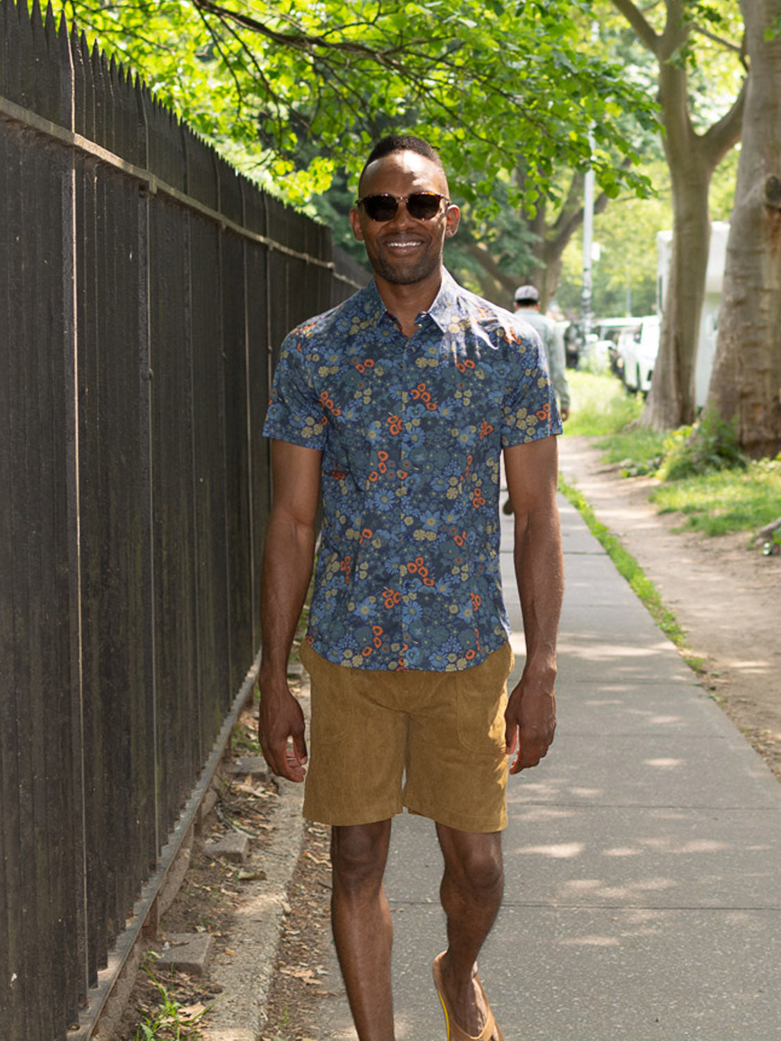 A man in sunglasses, a blue floral short-sleeve shirt, and tan ID Menswear Tunis Corduroy Shorts with an elastic waistband walks on a sunny sidewalk by a black fence and green trees.
