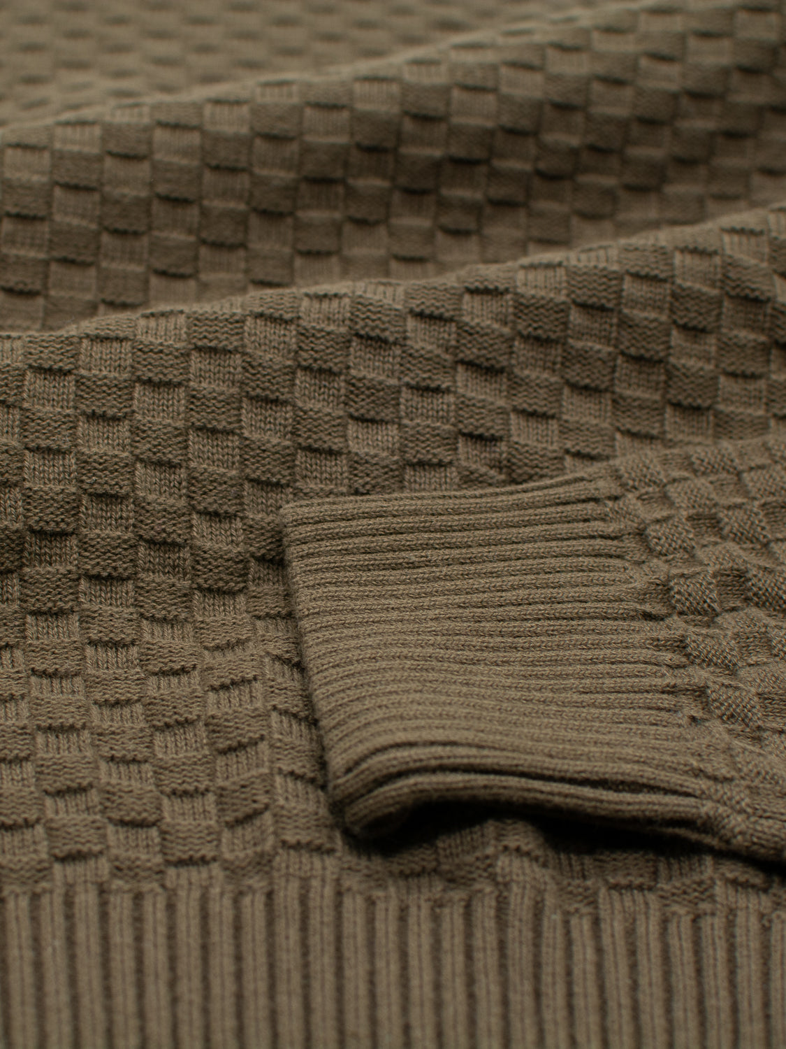 Close-up of the Turku Cotton Sweater by ID Menswear showcases its textured brown fabric with a grid-like knit pattern. The folded, ribbed section resembles a sweater cuff, giving it the appearance of a thick and warm checked cotton sweater.