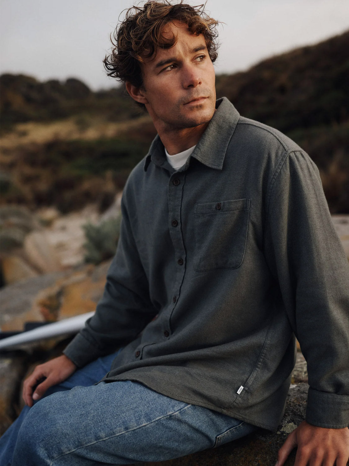A man with curly brown hair, wearing the Katin Twiller Flannel Button Down Shirt and blue jeans, sits thoughtfully on a rock outdoors with hills and natural scenery in the background.