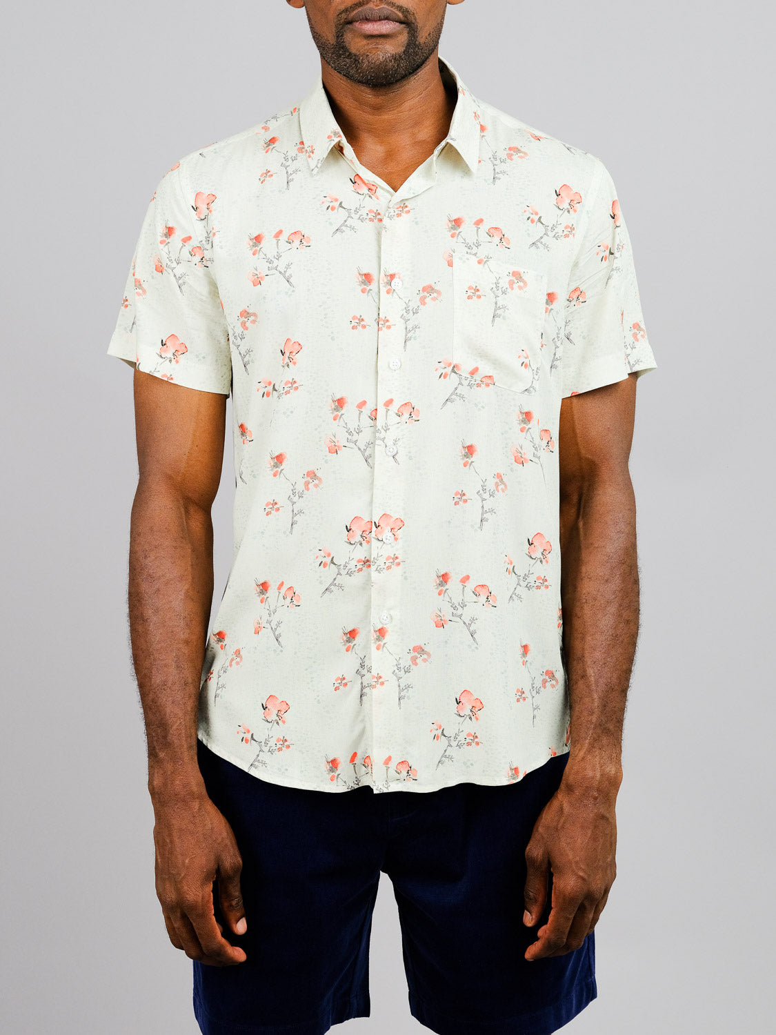 A man wearing the Ume Printed Rayon Shirt by ID Menswear, featuring vibrant pink and orange floral prints, paired with navy blue shorts, stands against a plain gray background.
