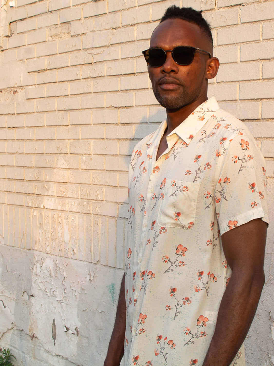 A man in the Ume Printed Rayon Shirt by ID Menswear—featuring a bold floral print and short sleeves—stands confidently before a pale brick wall, sunglasses on, gazing at the camera.