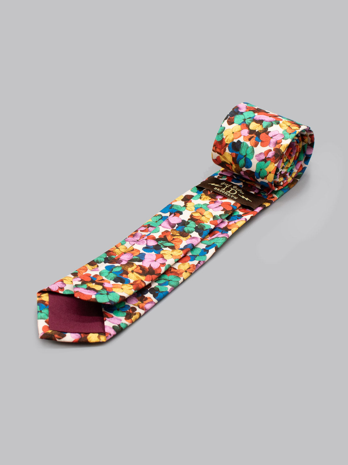 The ID Menswear Liberty of London Tana Lawn™ Hilary Tie features a vibrant multicolored floral pattern and is handmade in Brooklyn for a unique touch, shown rolled and unrolled on a plain gray background.