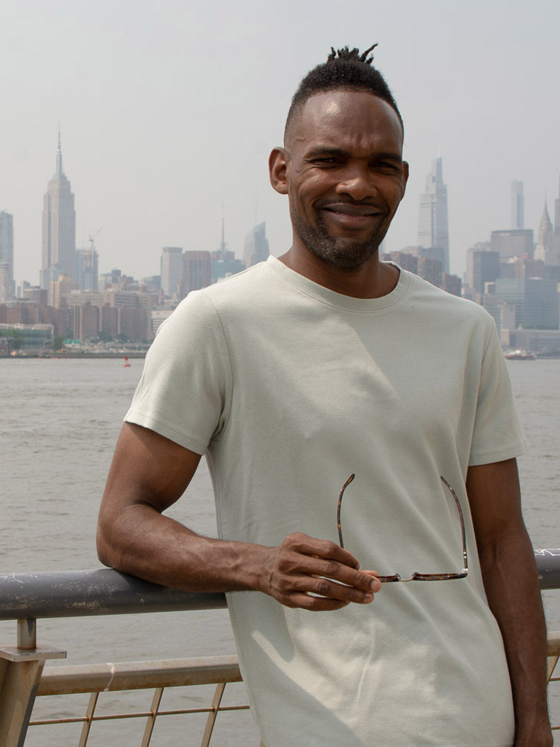 A man wearing the Urban Honeycomb Mesh Tee by ID Menswear smiles, holding sunglasses by a riverside railing. City skyscrapers, including the Empire State Building, rise in the hazy background across the water.