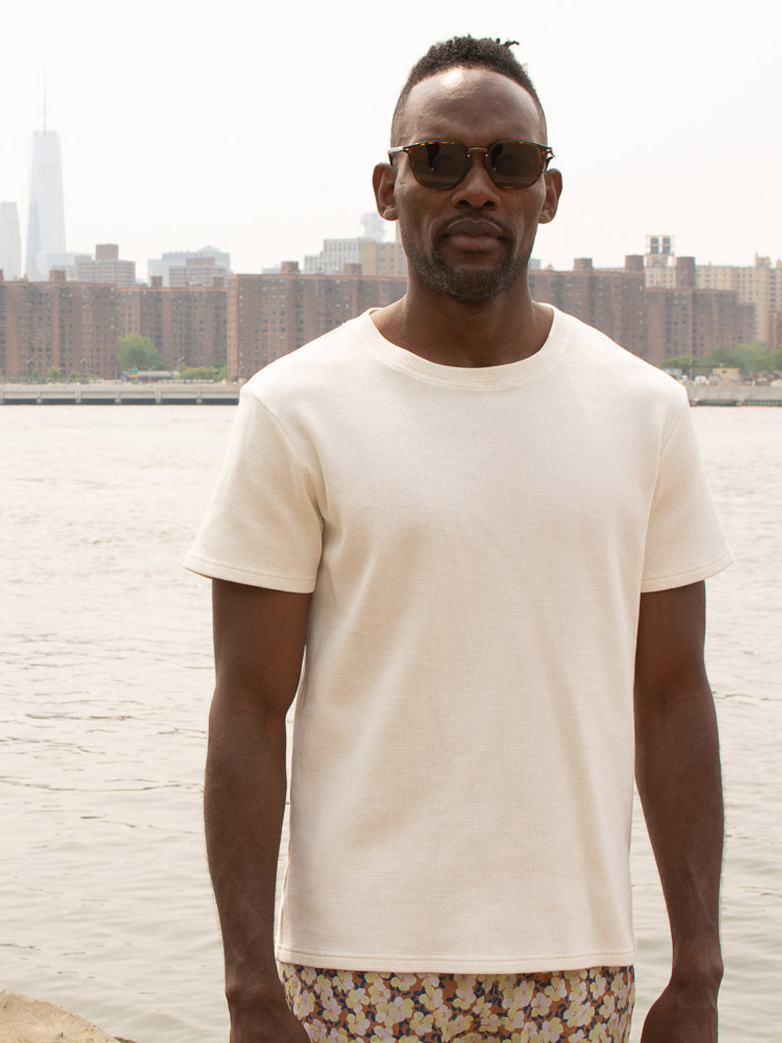A man in sunglasses and the ID Menswear Urban Waffle Knit Tee stands by the water, city buildings behind him on a bright day.