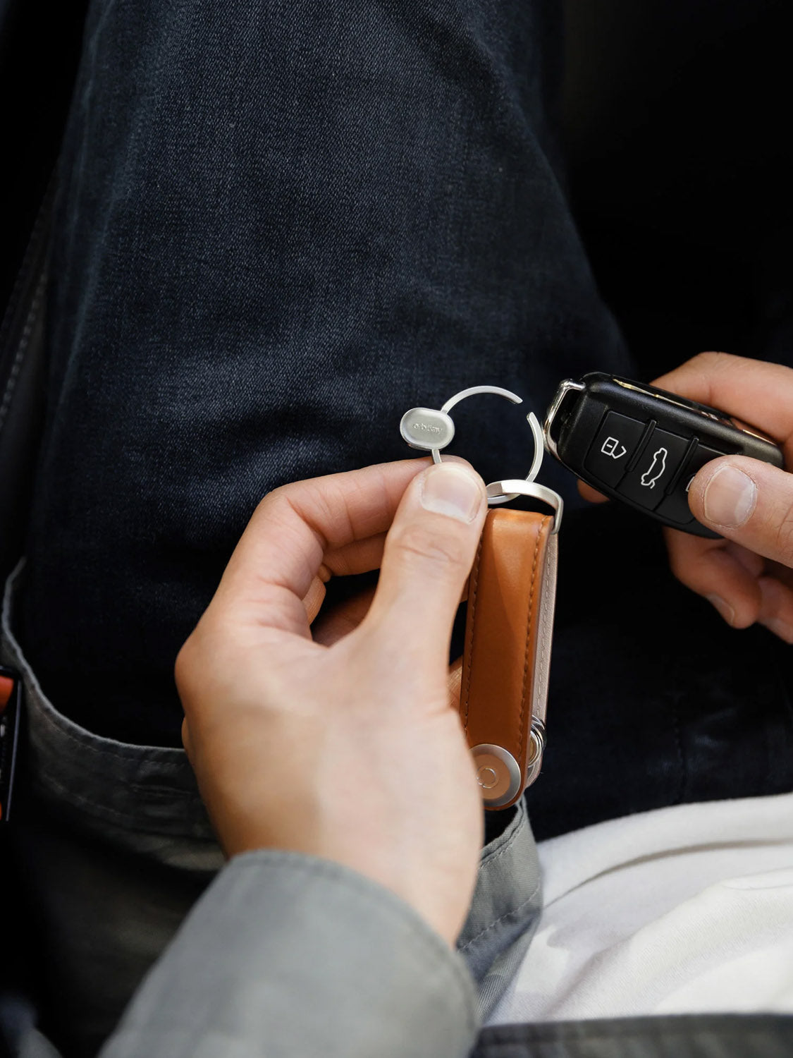 A person holds an Orbitkeys Key Ring V2 and a car key fob while sitting in dark jeans and a gray shirt.