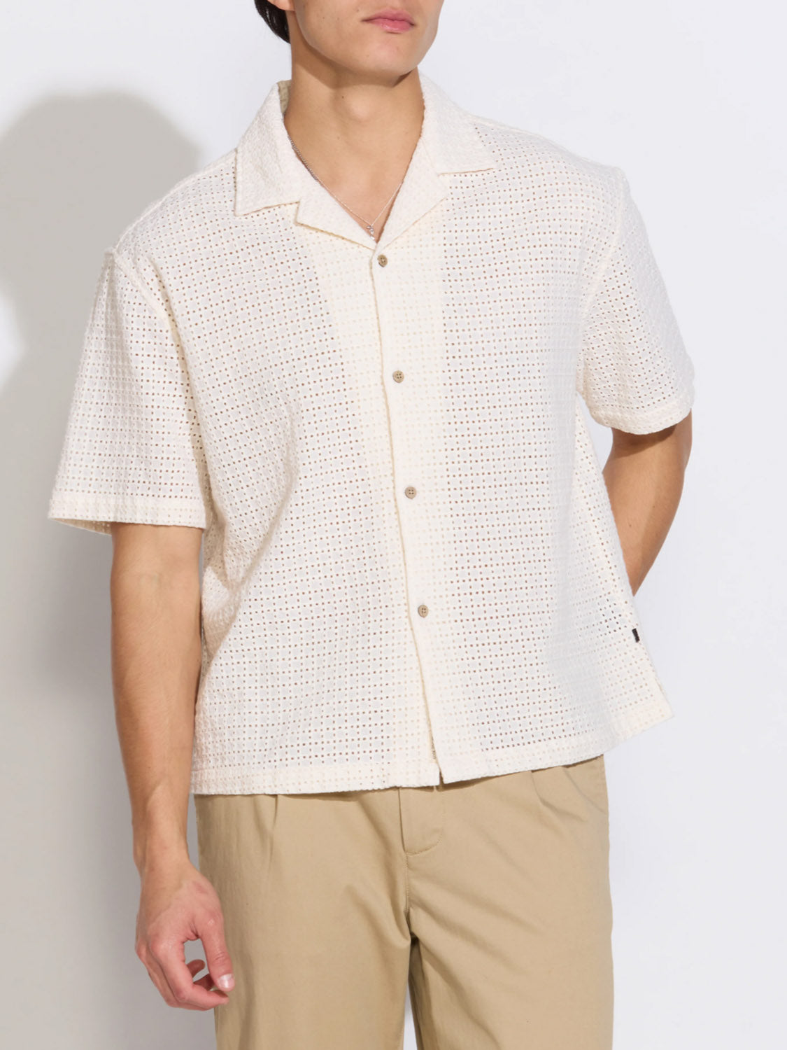 A person wears the Valencia Eyelet Resort Shirt by Civil Society—a cream-colored, cotton embroidered boxy shirt—paired with beige pants, standing against a plain white background.