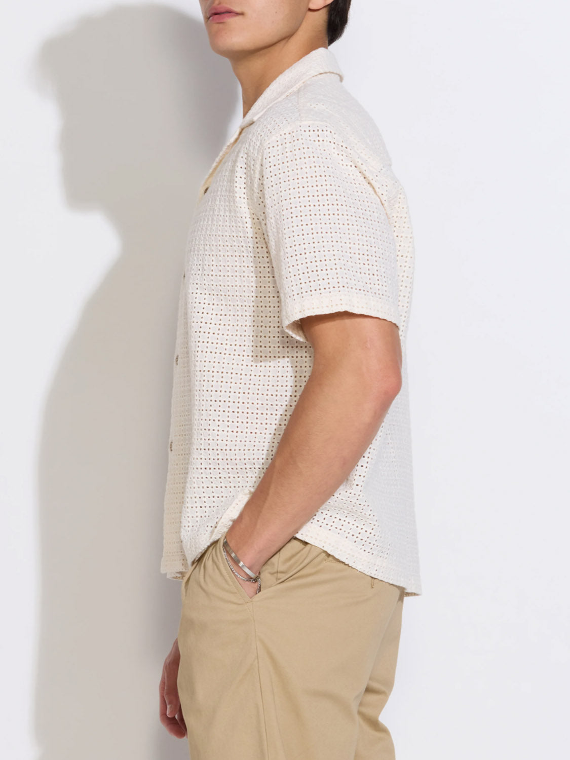 A person wears the Civil Society Valencia Eyelet Resort Shirt— a short-sleeve, cream-colored cotton embroidered top— with beige pants, standing sideways against a white background, their face partially out of frame.