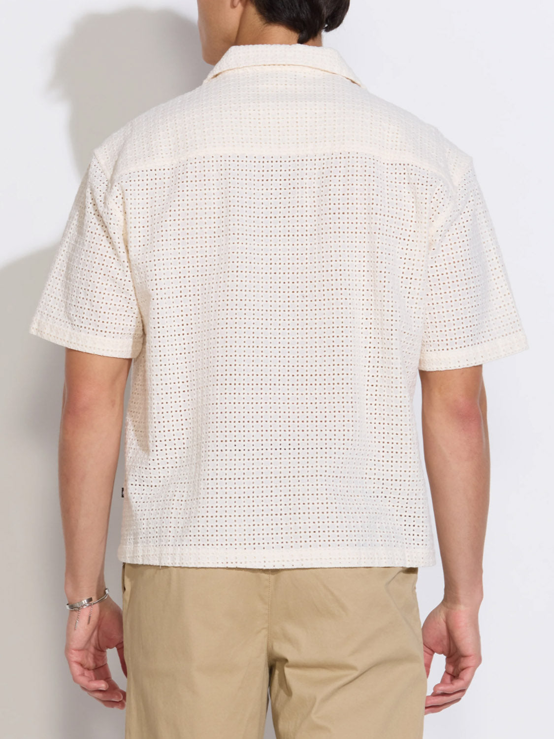 A person is seen from the back against a plain white background, wearing the Civil Society Valencia Eyelet Resort Shirt— a short-sleeve, cream cotton embroidered eyelet top with a boxy fit—paired with tan pants.