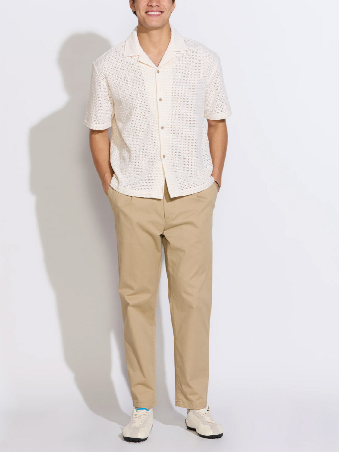 A person stands against a plain background, smiling with hands in pockets, wearing the Civil Society Valencia Eyelet Resort Shirt in white, paired with beige pants and white sneakers.