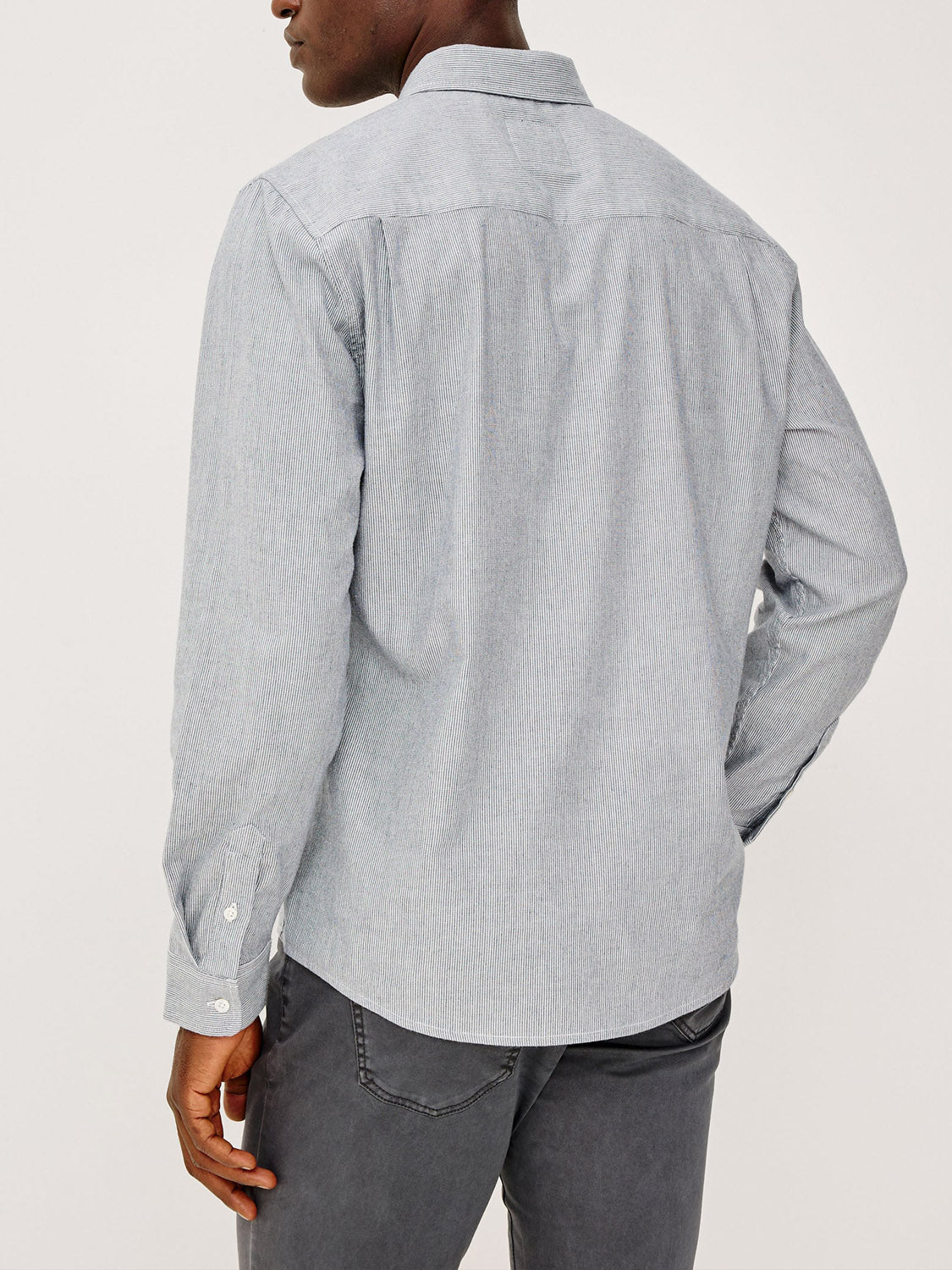 A man is seen from behind wearing the light gray Vance Shirt by Rails with gray pants. His left hand rests on his hip against a plain, light background, highlighting this cotton-twill button-up as perfect for everyday wear.