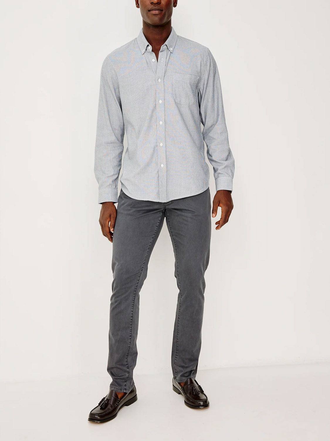 A man with a neutral expression stands against a white background, wearing the Rails Vance Shirt in light blue cotton-twill, paired with gray slim-fit pants and black loafers. His arms rest relaxed by his sides.