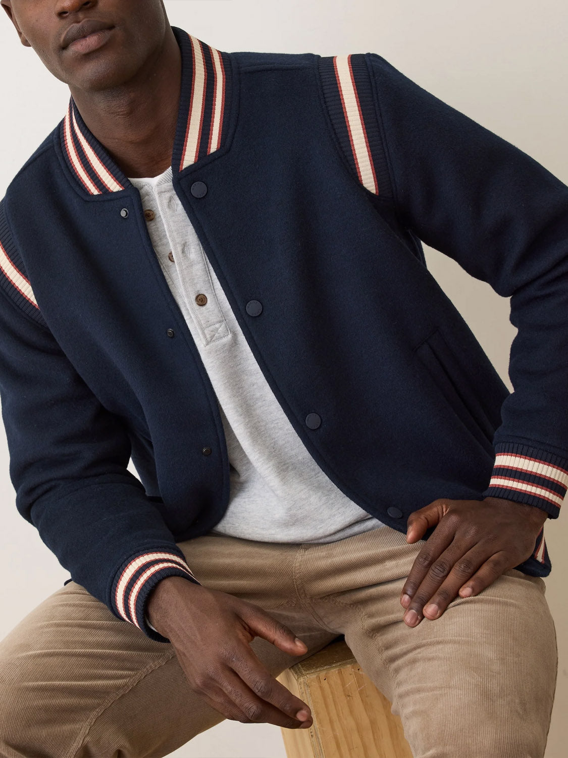 A man sits on a wooden stool wearing the Marine Layer Fields Wool Varsity Jacket, a classic-fit navy piece with red and white striped trim, paired with a light gray henley and tan corduroy pants. His head is slightly turned and his hand rests on his knee.